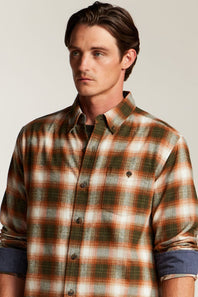 A man wearing a long-sleeve button-down plaid shirt in green, orange, and white, with rolled-up sleeves showing a blue undershirt underneath, set against a plain light gray background.