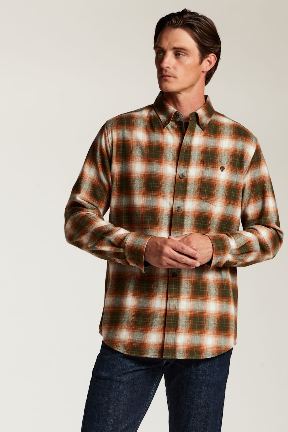 A man wearing a long-sleeve button-up plaid flannel shirt in brown, orange, and white, paired with dark blue jeans, photographed against a light gray studio background.