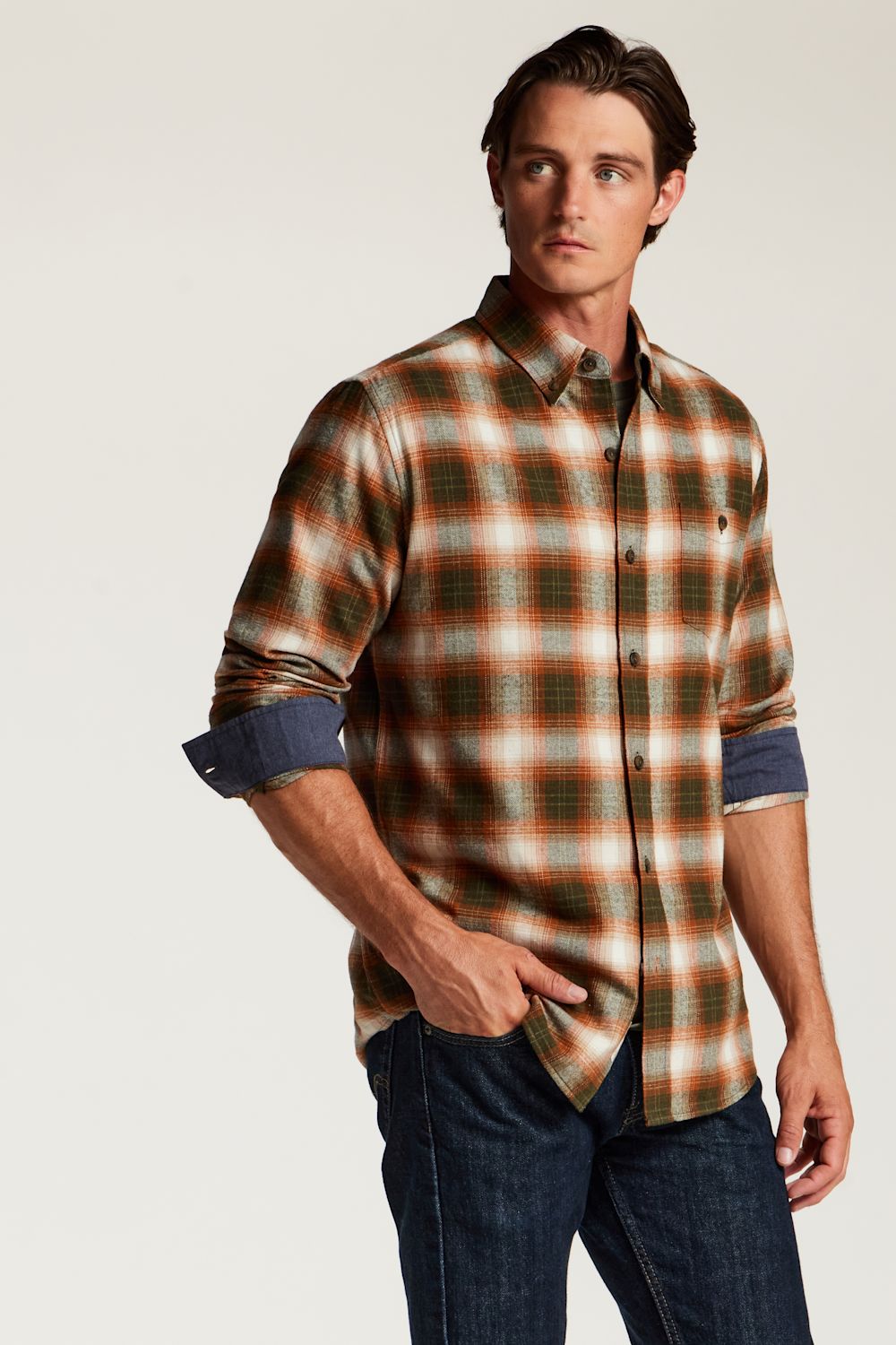 A man wearing a long-sleeve button-down plaid shirt in brown, orange, and green tones with rolled-up sleeves showing a blue undershirt, paired with dark blue jeans, against a light gray studio background.