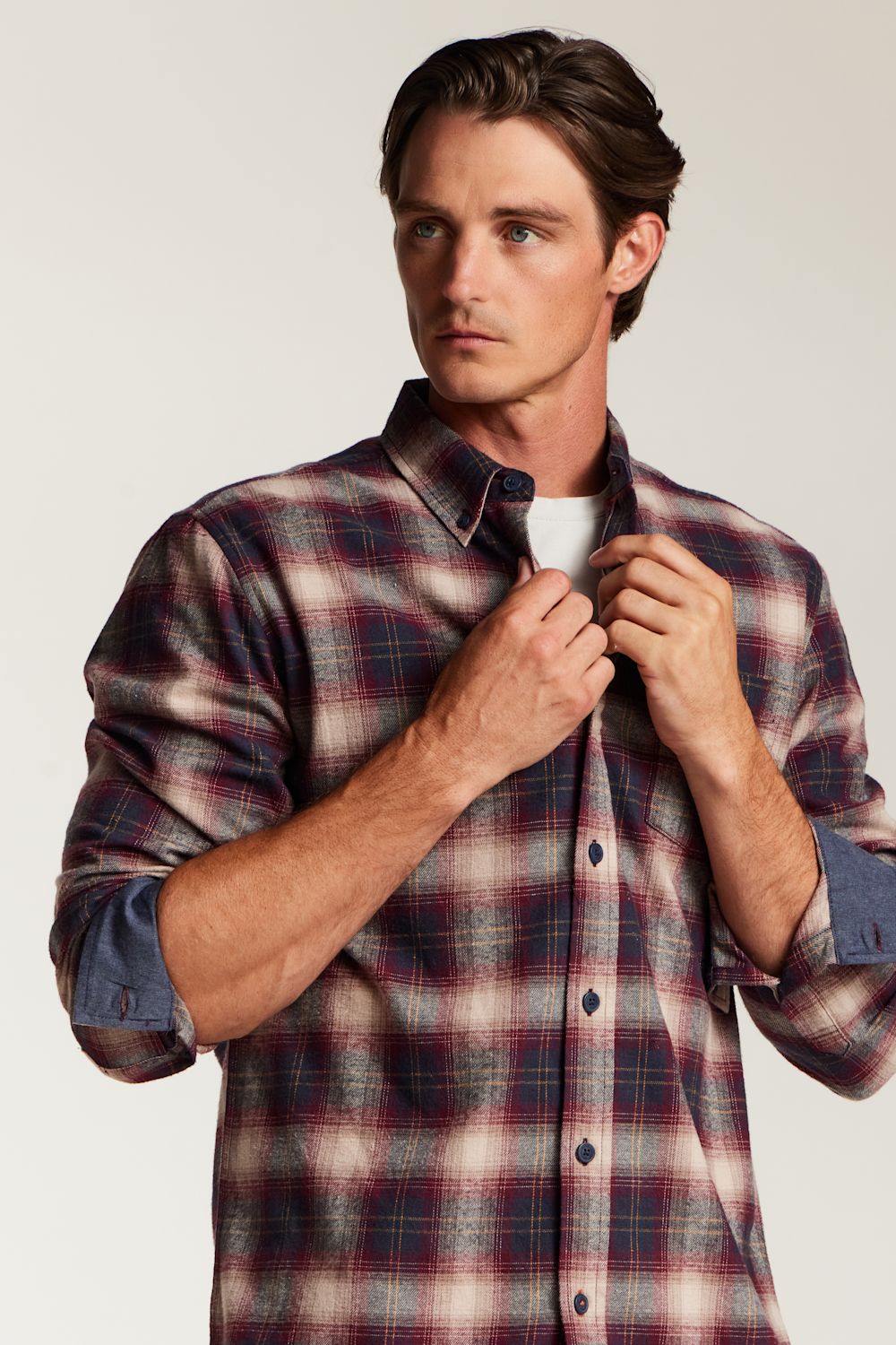 A man wearing a long-sleeve plaid button-up shirt in red, navy, and beige tones, adjusting the collar of the shirt against a plain light background.