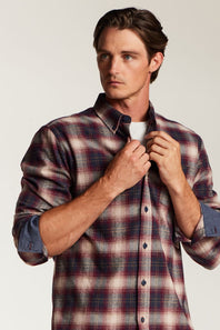 A man wearing a long-sleeve plaid button-up shirt in red, navy, and beige tones, adjusting the collar of the shirt against a plain light background.