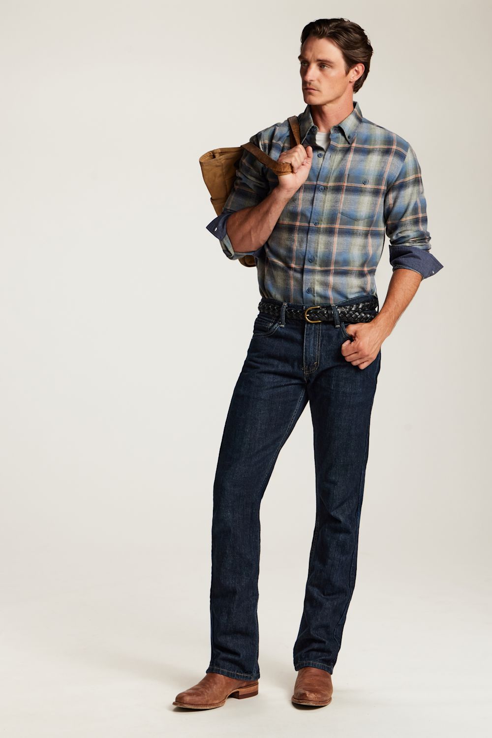 A man modeling a blue and gray plaid button-down shirt, dark blue jeans, a black belt, and brown leather boots, holding a brown leather shoulder bag against a plain off-white studio background.