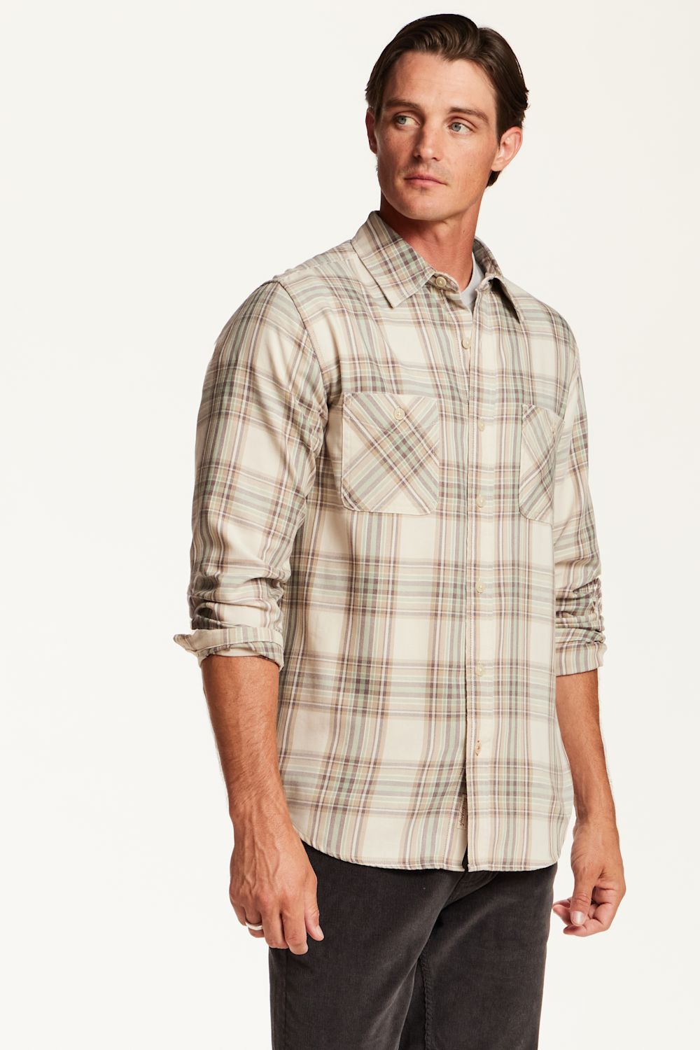 A man wearing a long-sleeve plaid button-up shirt in beige, brown, and white, paired with dark gray pants, photographed against a clean white studio background.