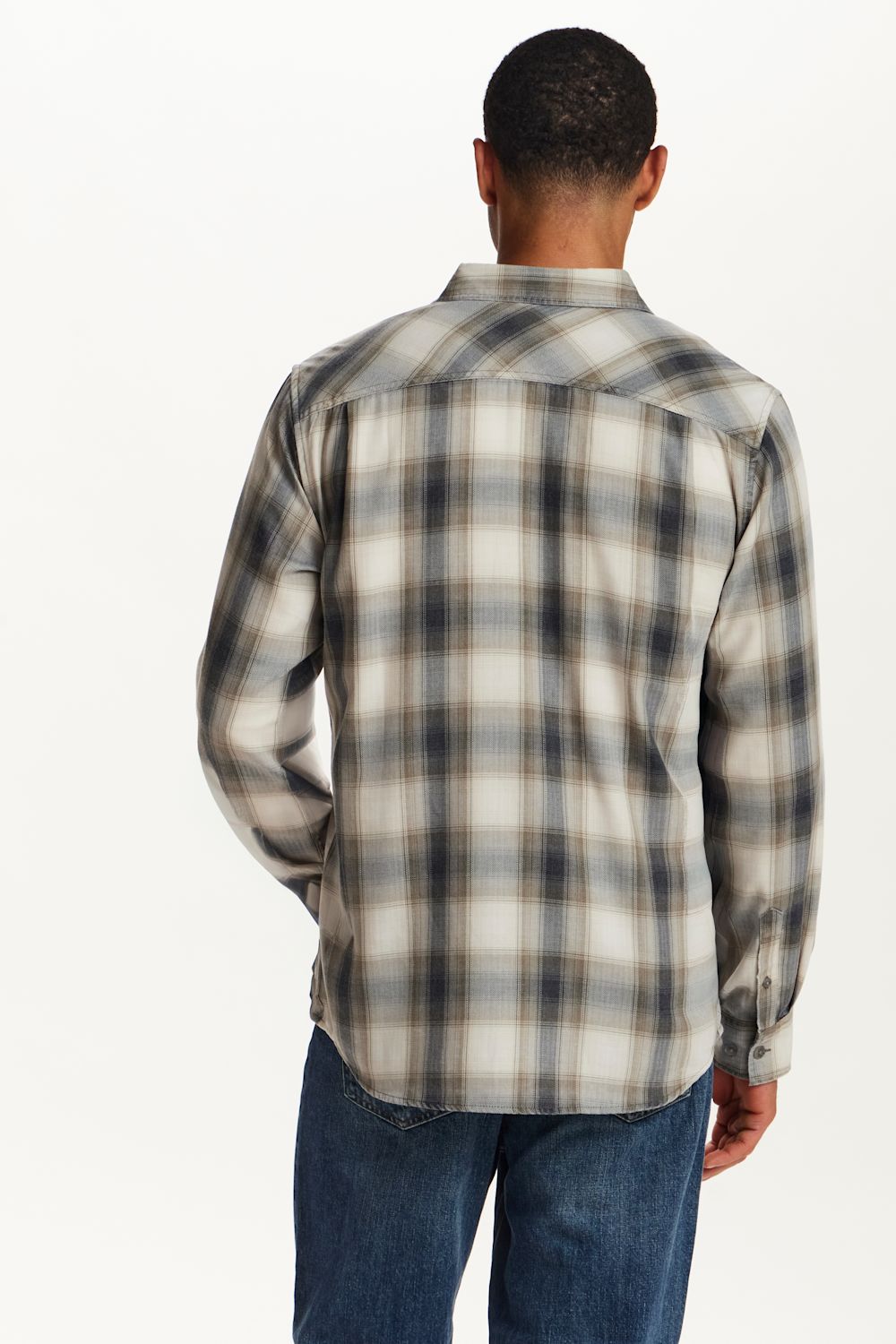 A man viewed from behind wearing a long-sleeve plaid button-up shirt in beige, gray, and black, paired with blue jeans, against a plain white background.