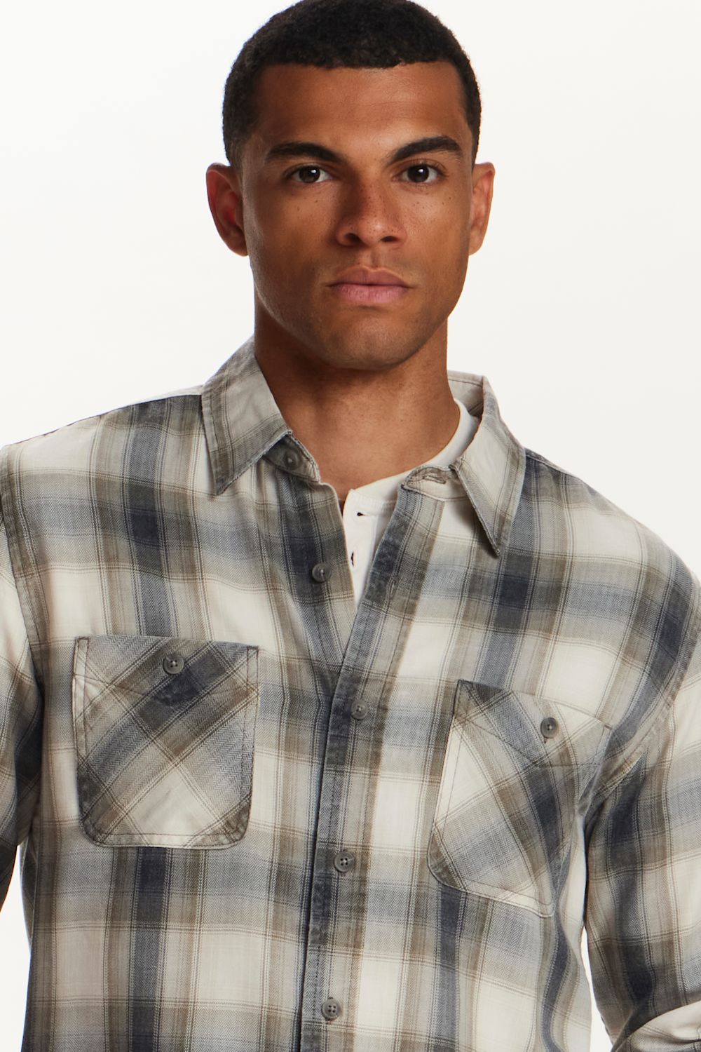A man wearing a long-sleeve plaid button-up shirt in beige, gray, and white, layered over a white undershirt, against a plain white background.