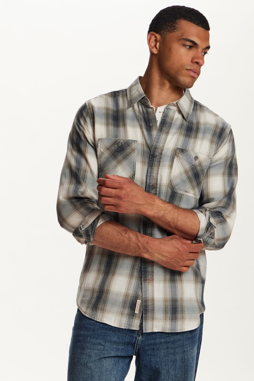 A man wearing a long-sleeve plaid button-up shirt in gray, white, and black, paired with blue jeans, photographed against a clean white studio background.