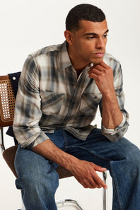 A man sitting on a chair wearing a gray and white plaid button-down shirt with blue jeans, posed against a clean white studio background.