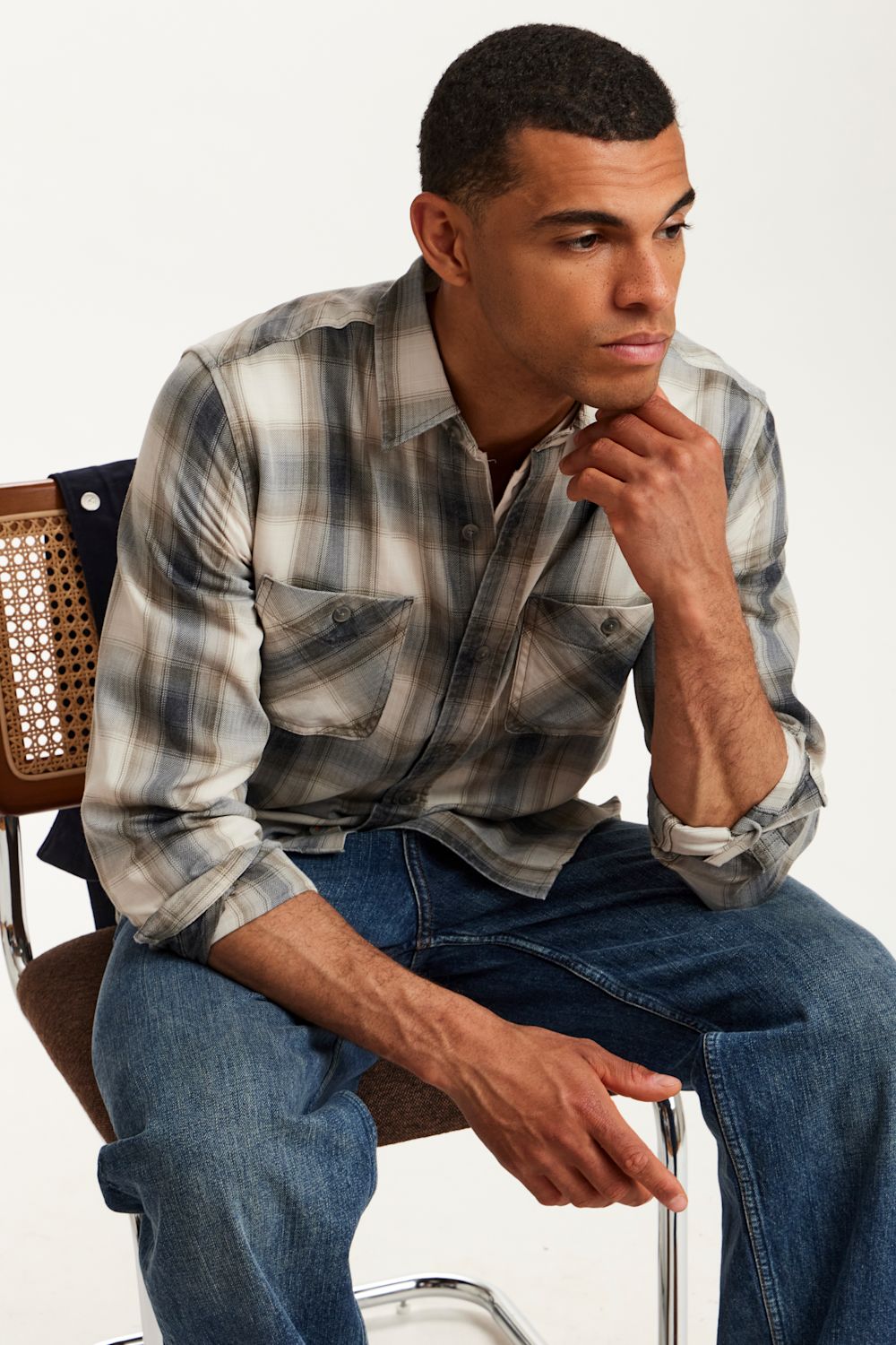A man sitting on a chair wearing a gray and white plaid button-down shirt with blue jeans, posed against a clean white studio background.