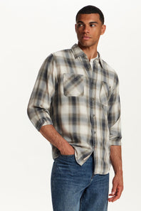 A man wearing a long-sleeve plaid button-up shirt in beige, gray, and white, paired with blue jeans, photographed against a clean white studio background.