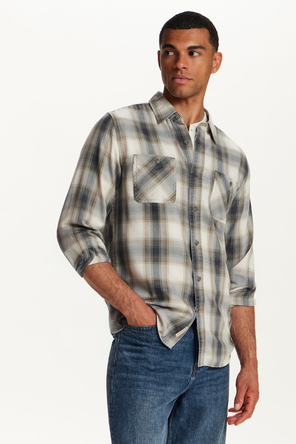 A man wearing a long-sleeve plaid button-up shirt in beige, gray, and white, paired with blue jeans, photographed against a clean white studio background.