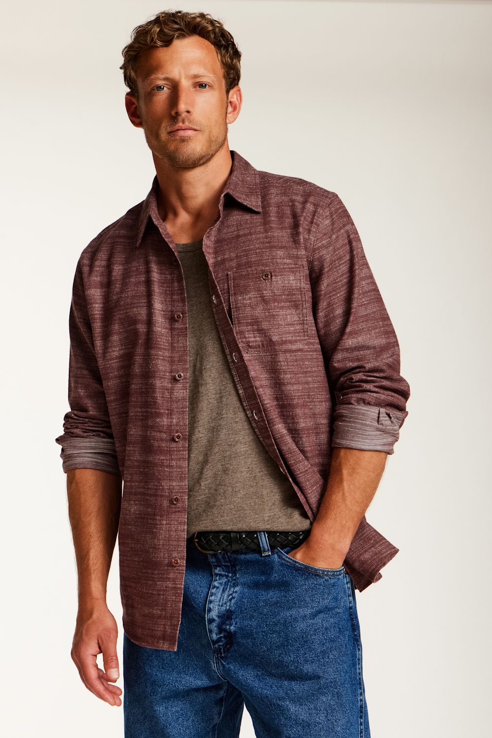 A man wearing a textured, heathered burgundy button-down shirt over a gray t-shirt and blue jeans, standing against a plain light background.