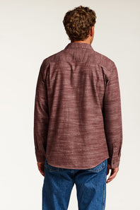 A man viewed from behind wearing a textured, heathered burgundy long-sleeve button-down shirt and blue jeans against a plain white studio background.