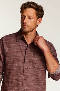 A man wearing a textured, heathered burgundy button-down shirt with rolled-up sleeves, posing against a light off-white background.