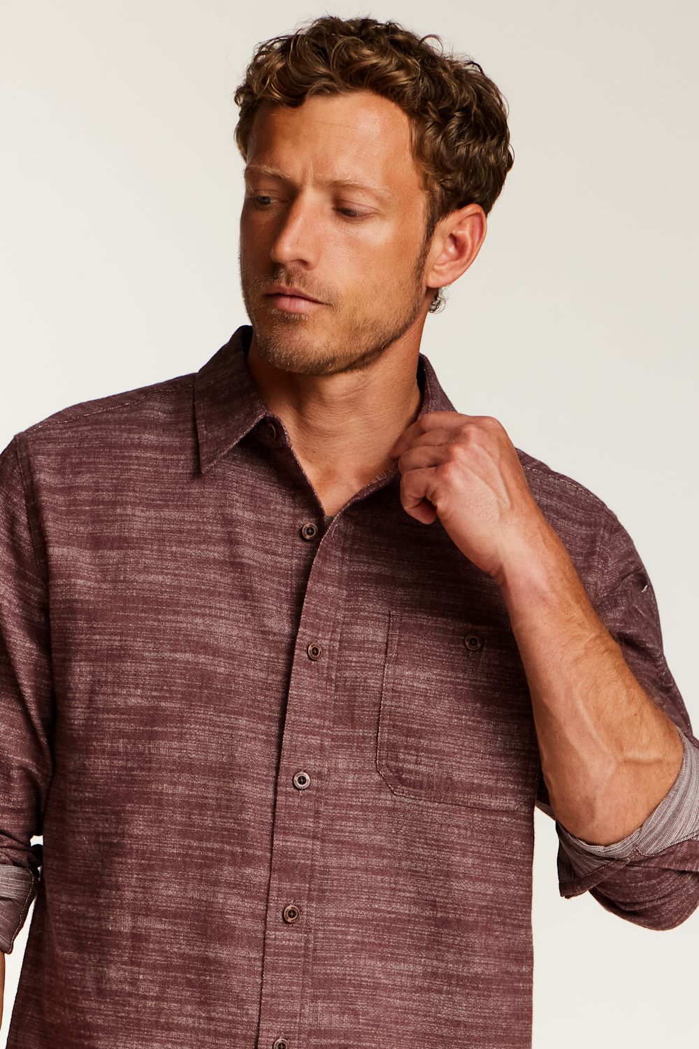 A man wearing a textured, heathered burgundy button-down shirt with rolled-up sleeves, posing against a light off-white background.