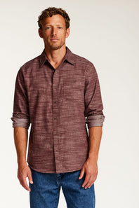 A man wearing a textured, heathered burgundy button-down shirt with rolled-up sleeves and blue jeans, standing against a plain off-white studio background.