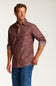 A man wearing a textured, heathered burgundy button-down shirt with rolled-up sleeves and blue jeans, standing against a plain light gray studio background.