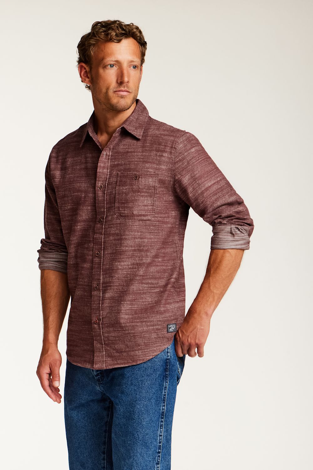 A man wearing a textured, heathered burgundy button-down shirt with rolled-up sleeves and blue jeans, standing against a plain light gray studio background.