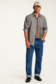A man modeling a gray long-sleeve button-down shirt over a brown t-shirt, paired with blue jeans, black shoes, and a beige baseball cap, against a plain white studio background.