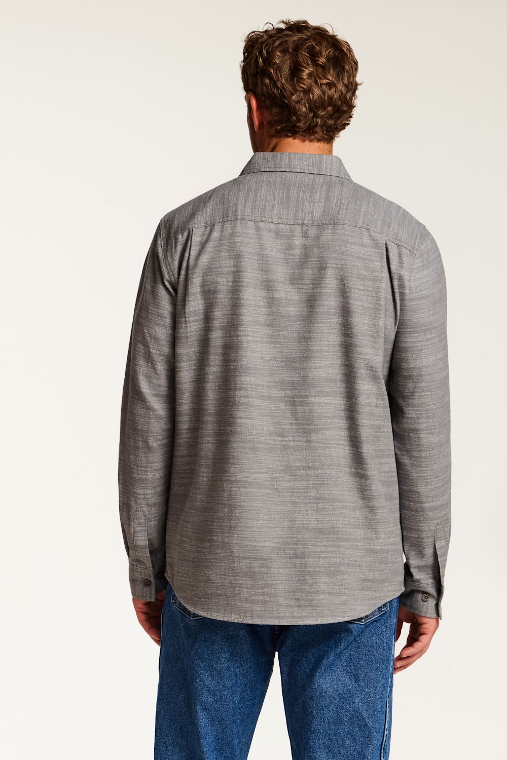 A man viewed from behind wearing a gray textured long-sleeve button-up shirt and blue jeans against a plain white studio background.
