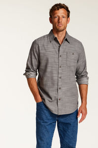 A man wearing a gray long-sleeve button-down shirt with rolled-up sleeves and blue jeans, standing against a plain light background.
