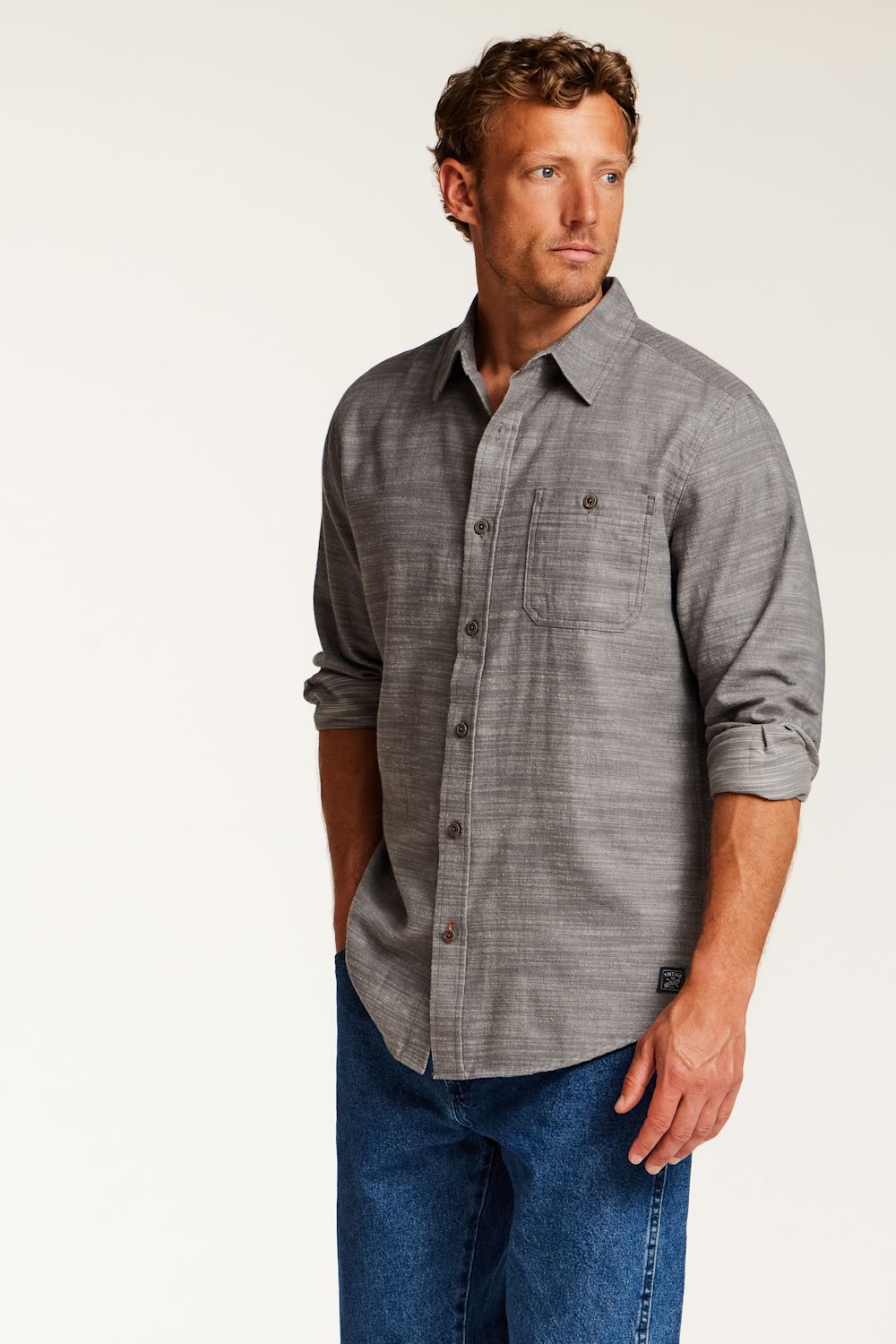 A man wearing a gray long-sleeve button-down shirt with rolled-up sleeves and blue jeans, standing against a plain light background.