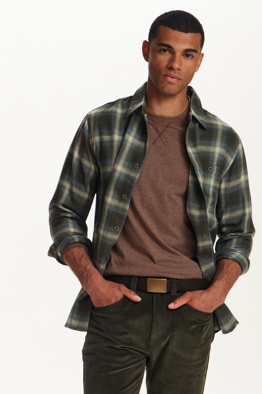 A man wearing a green, blue, and white plaid button-down shirt over a brown crewneck t-shirt, paired with dark green pants and a gold-tone belt, against a plain white background.