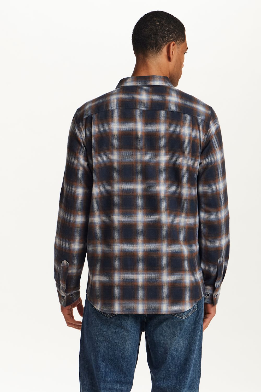 A man viewed from the back wearing a long-sleeve plaid button-up shirt in dark navy, brown, and beige tones, paired with blue jeans, against a plain white background.