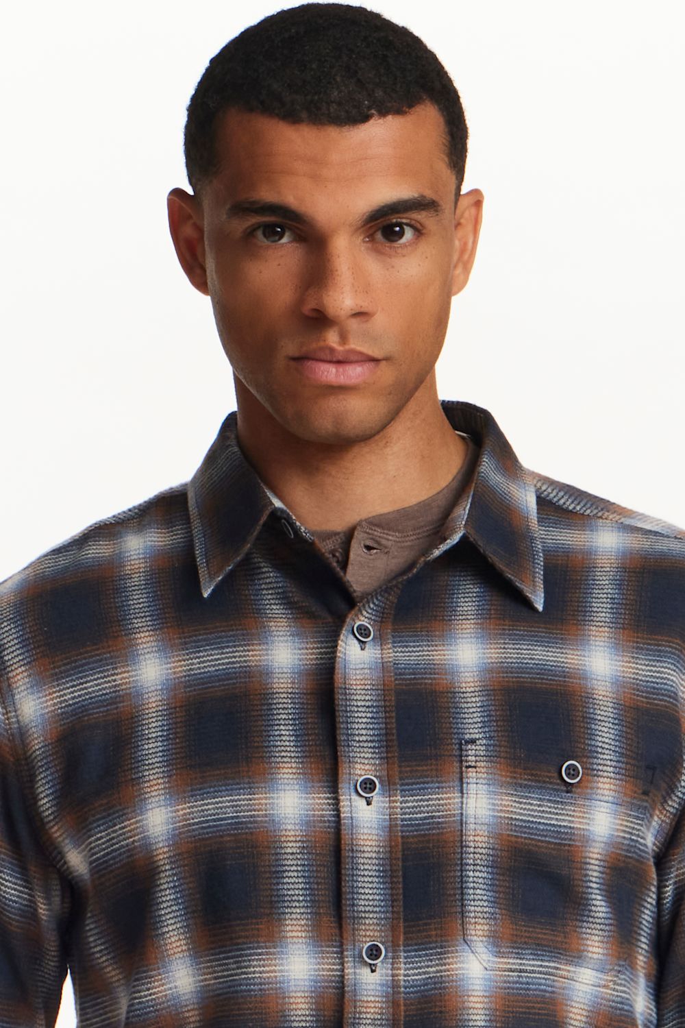 A man wearing a navy blue and brown plaid button-down shirt over a brown undershirt, photographed against a clean white background.