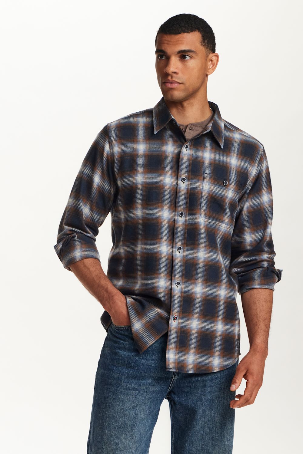 A man wearing a long-sleeve plaid button-up shirt in navy, brown, and white, paired with blue jeans, photographed against a clean white studio background.