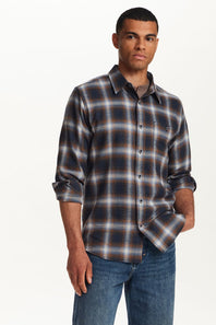 A man wearing a long-sleeve button-up plaid shirt in dark blue, brown, and white, paired with blue jeans, photographed against a clean white background.