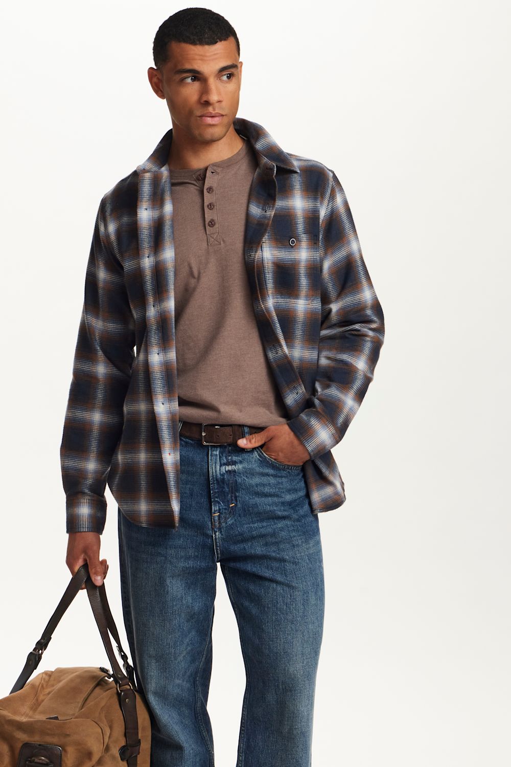 A man modeling a dark brown and navy plaid overshirt over a brown henley shirt, paired with blue jeans and holding a tan duffel bag, set against a plain white studio background.