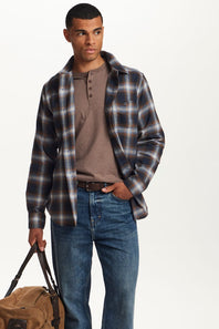A man modeling a dark brown and navy plaid overshirt over a brown henley shirt, paired with blue jeans and holding a tan duffel bag, set against a plain white studio background.