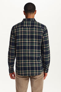 A man viewed from the back wearing a long-sleeve plaid button-up shirt in navy, green, and beige, paired with tan pants, against a plain white background.