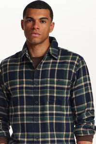 A man wearing a long-sleeve button-up plaid flannel shirt in navy blue, forest green, and beige, layered over a dark brown t-shirt, against a plain white background.