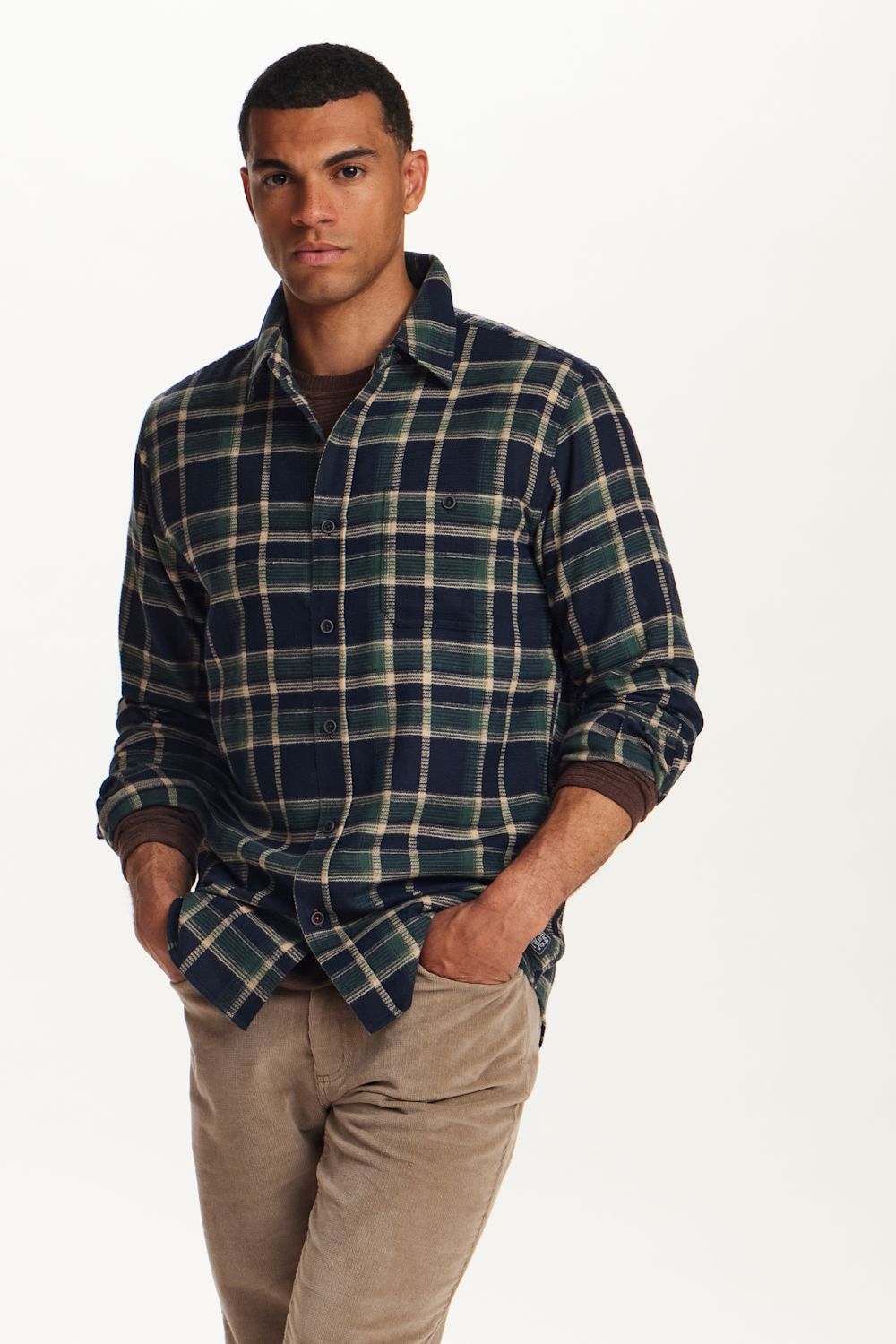 A man wearing a navy blue and green plaid button-down flannel shirt with beige pants, standing against a plain white studio background.