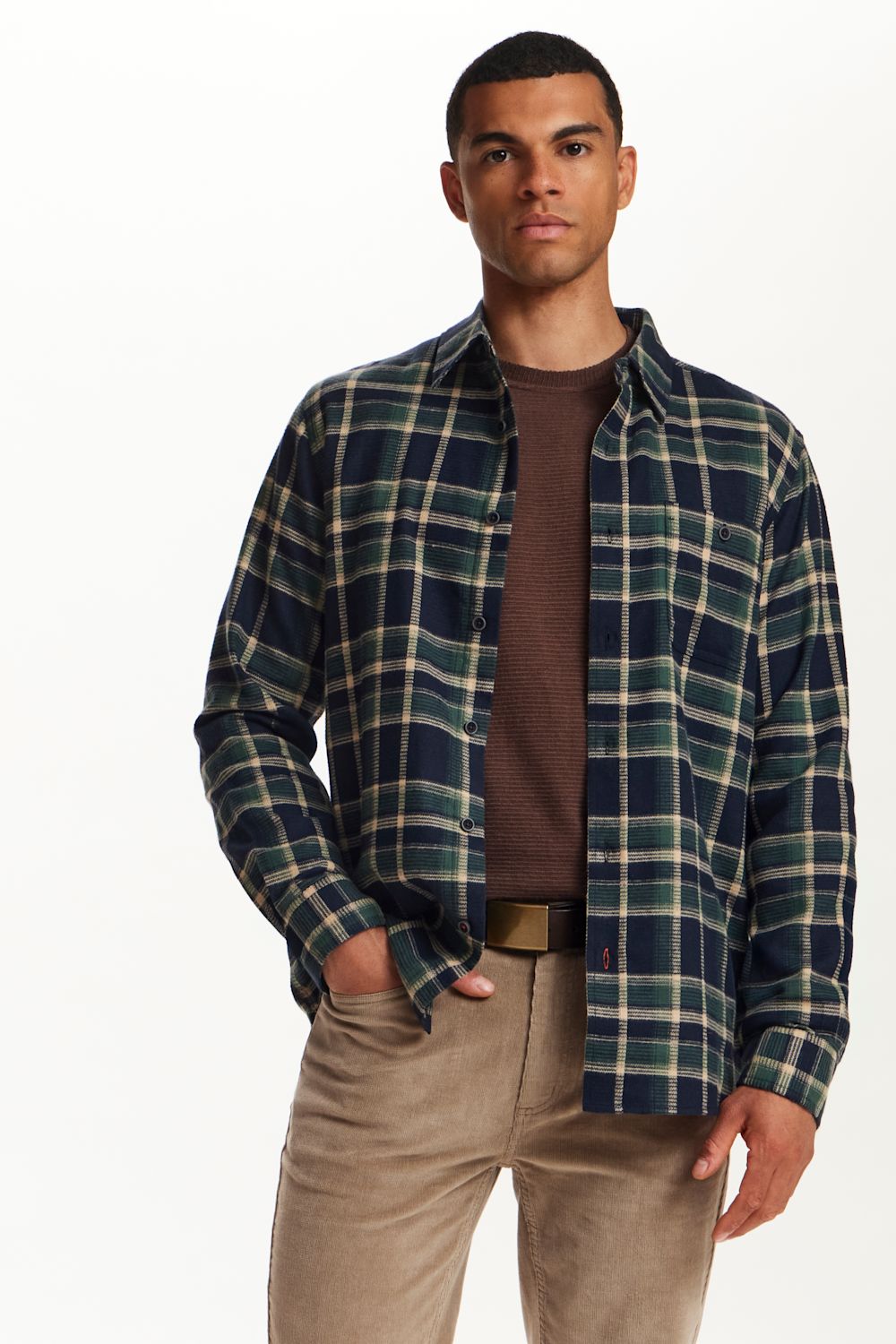 A man wearing a navy blue and green plaid button-down shirt over a brown ribbed t-shirt, paired with beige trousers, against a plain white background.