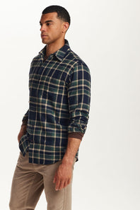 A man wearing a navy blue and green plaid long-sleeve button-down shirt with brown cuffs, paired with beige pants, photographed against a plain white studio background.
