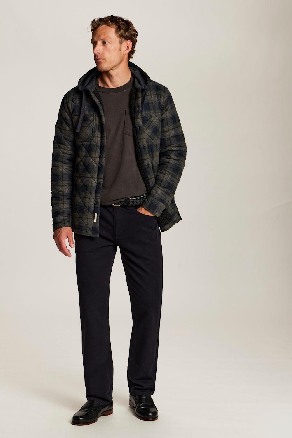 A man modeling a dark green and black plaid hooded jacket over a dark gray t-shirt, black trousers, and black leather shoes, standing against a light gray studio background.