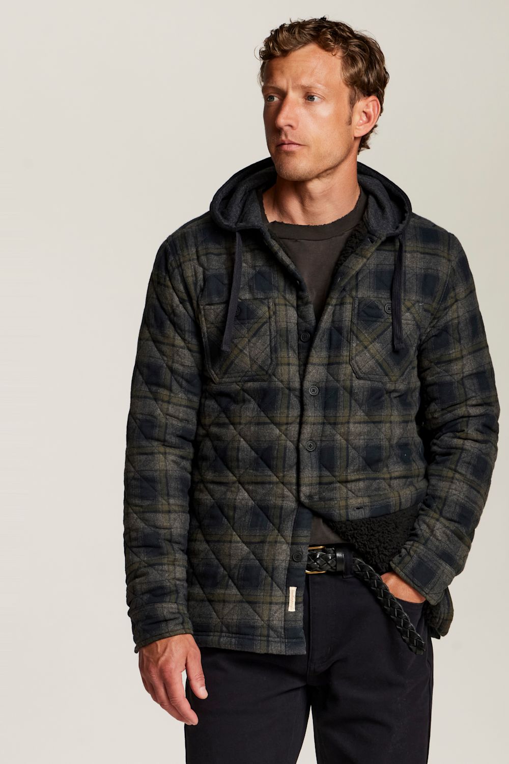 A man wearing a dark navy and olive green quilted hooded jacket with a subtle plaid pattern, styled over a dark t-shirt and black trousers, photographed against a neutral light gray studio background.