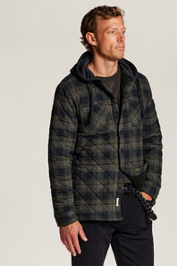 A man wearing a dark navy and black plaid quilted jacket with a hood, styled over a dark t-shirt and black pants, photographed against a light gray studio background.
