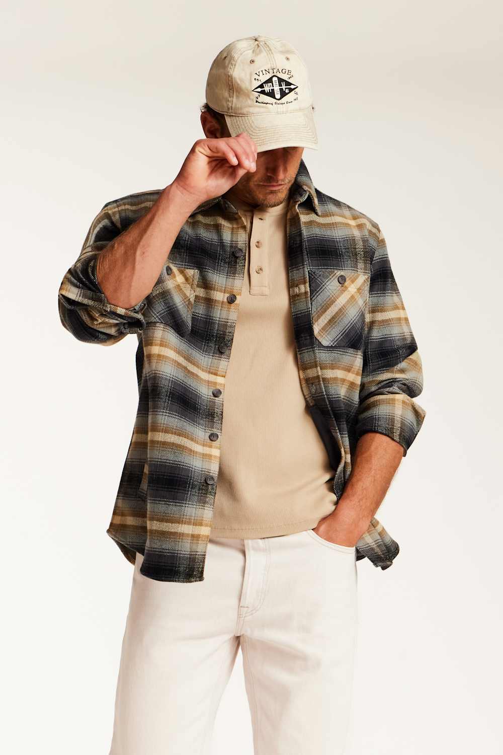 A man wearing a beige baseball cap with a black graphic, a brown and black plaid flannel shirt over a beige henley t-shirt, and off-white pants, posed against a clean white studio background.