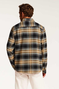 A man viewed from behind wearing a long-sleeve plaid button-up shirt in black, beige, and white, paired with off-white pants, against a plain white studio background.