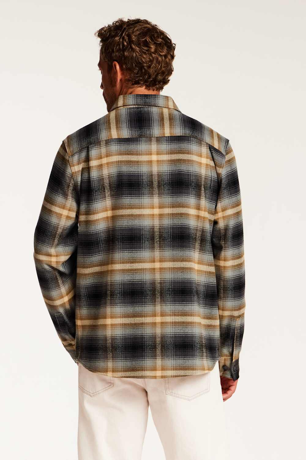 A man viewed from behind wearing a long-sleeve plaid button-up shirt in black, beige, and white, paired with off-white pants, against a plain white studio background.