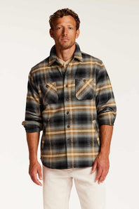 A man wearing a long-sleeve button-up plaid flannel shirt in black, beige, and brown tones, paired with off-white pants, against a plain white studio background.