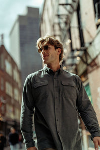 A man wearing a dark gray long-sleeve button-up shirt and sunglasses walking down an urban street.