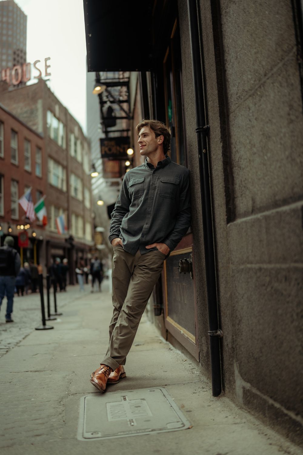 A man wearing a dark gray button-down shirt, khaki pants, and brown leather shoes leans against a dark stone building on a city sidewalk.