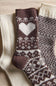 Three knitted socks arranged on a beige fabric background: one cream sock, one dark brown sock with a large white heart and white Fair Isle pattern, and one dark brown and cream patterned sock.
