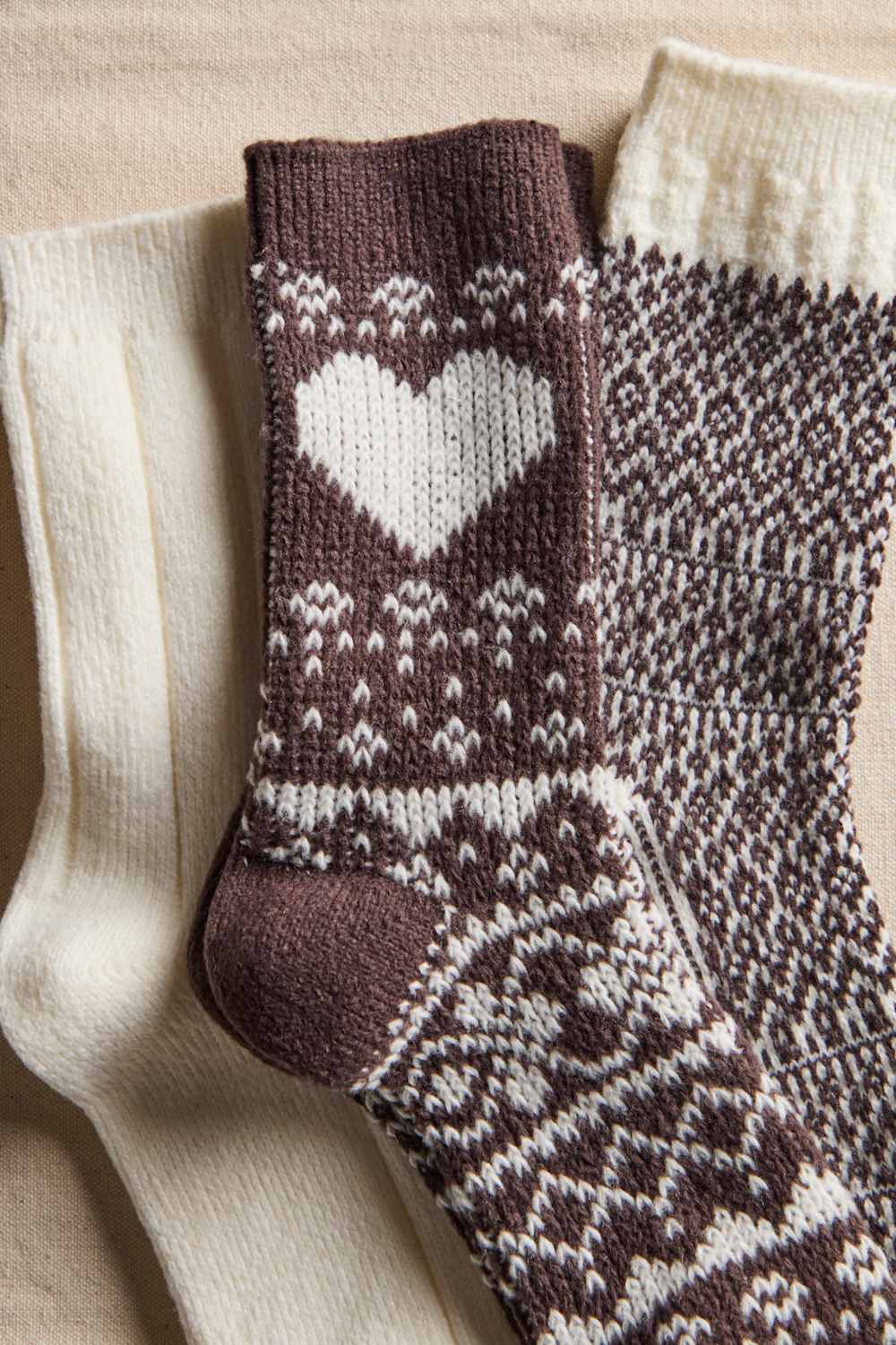 Three knitted socks arranged on a beige fabric background: one cream sock, one dark brown sock with a large white heart and white Fair Isle pattern, and one dark brown and cream patterned sock.
