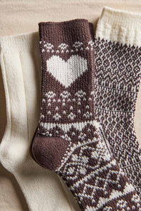 Three knitted socks arranged on a beige fabric background: one cream sock, one dark brown sock with a large white heart and white Fair Isle pattern, and one dark brown and cream patterned sock.