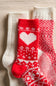 Three knit socks arranged on a beige fabric background: one white sock, one red-and-white heart pattern sock, and one red-and-white Nordic pattern sock.